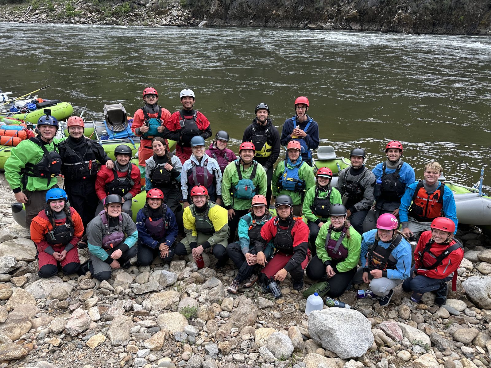 Roughly thirty river guides in drysuits and helmets gathered on a rocky beach beside rafts on the Salmon River in Idaho.